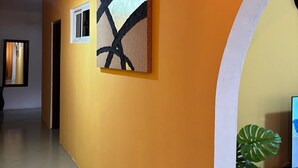 Un pasillo con pared amarilla, dos cuadros abstractos, una planta en maceta y una puerta que da a otra habitación.