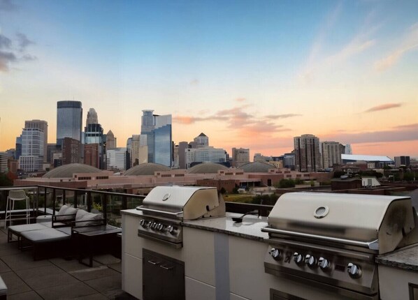 A rooftop patio with a grill and seating area overlooking a city skyline at sunset.