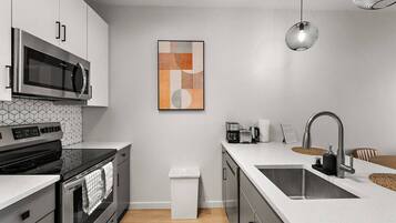 A modern kitchen with a stainless steel microwave, oven, and sink. There is a coffee maker, a trash bin, and a hanging light fixture.