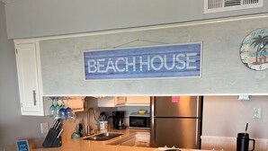 A kitchen with a 'BEACHHOUSE' sign, two bar stools, a microwave, a coffee maker, and a refrigerator.