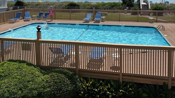 A rectangular swimming pool with a wooden deck and lounge chairs, surrounded by a wooden fence.