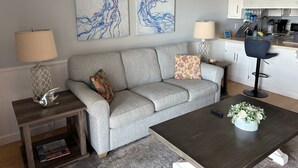 A living room with a grey sofa, two side tables with lamps, a coffee table with a plant, and a kitchen area with a 'BEACH' sign.