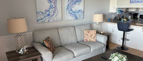 A living room with a grey sofa, two side tables with lamps, a coffee table with a plant, and a kitchen area with a 'BEACH' sign.