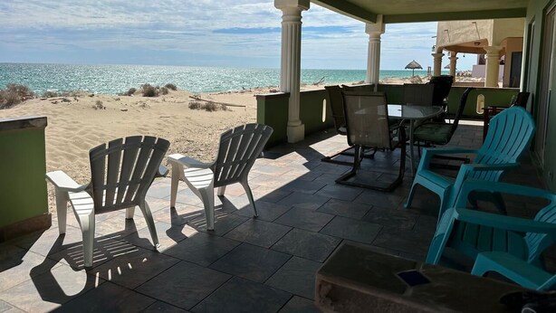 A covered outdoor seating area with chairs and tables, overlooking a beach with the ocean in the background.