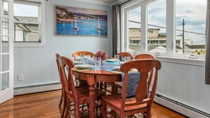A dining area with a table set for six, a painting of a coastal scene, and a view of buildings through large windows. - Hampton Beach, Free Parking, Sunsets (Hampton)