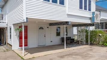 A white two-story house with a covered porch, a red door, and a small outdoor seating area.