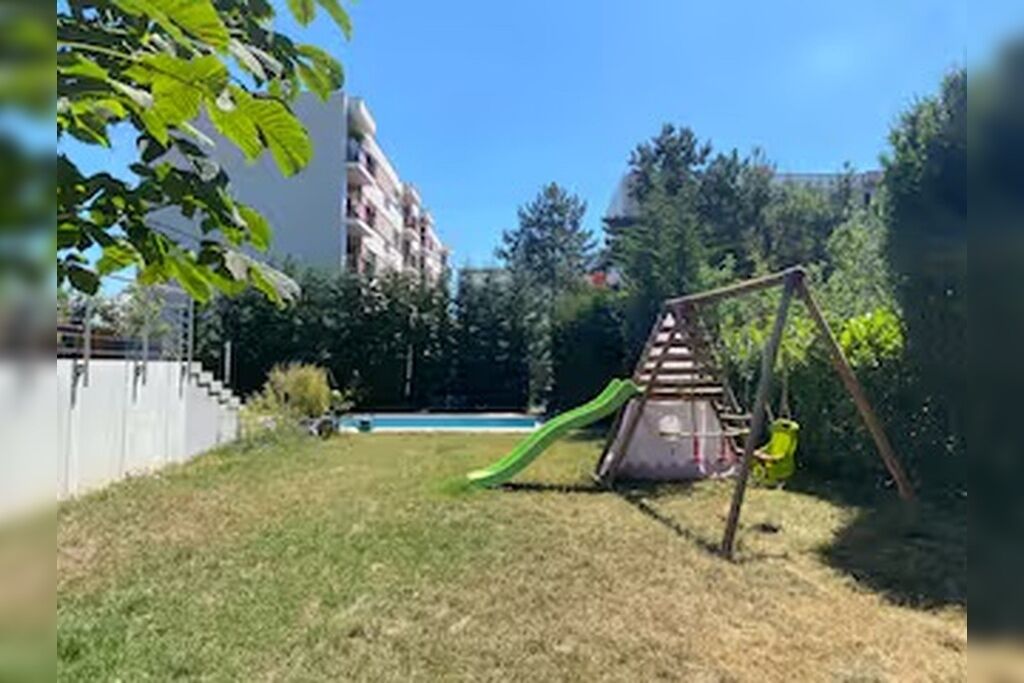 Un terrain de jeux avec un toboggan et des balançoires, entouré d’herbe et d’arbres, avec des bâtiments en arrière-plan.