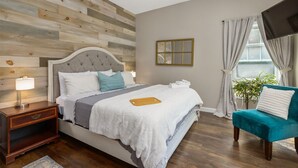 Un dormitorio con cabecera de madera, cama con ropa blanca, mesita de noche con lámpara, sillón azul y planta en maceta junto a la ventana.