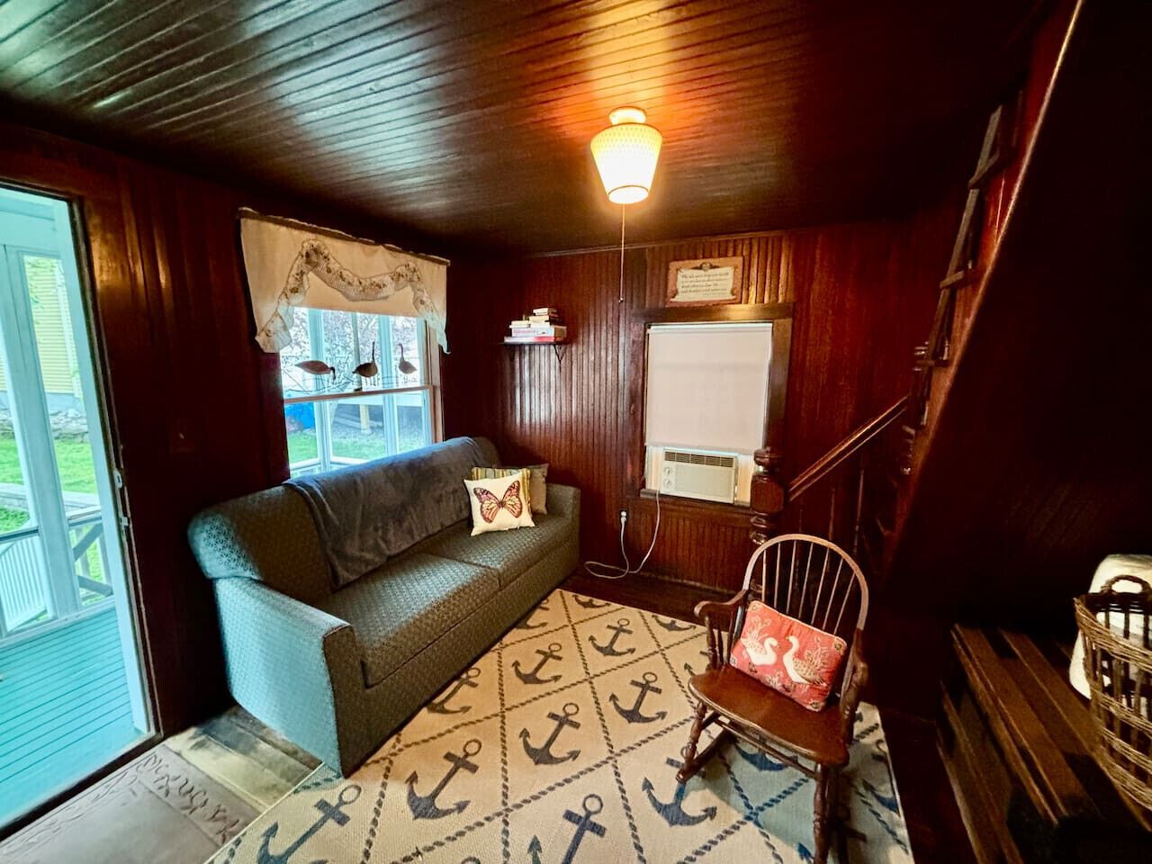A cozy room with a wooden ceiling, a sofa with a cushion, a patterned rug, a chair with a cushion, and a window with a curtain.