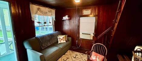 A cozy room with a wooden ceiling, a sofa with a cushion, a patterned rug, a chair with a cushion, and a window with a curtain.