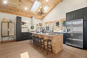 Eine moderne Küche mit einer zentralen Kochinsel, Barhocker, einem großen Kühlschrank und einer Holzbalkendecke mit Oberlichtern.