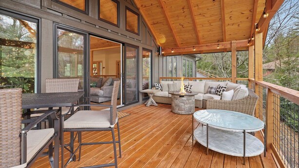 A covered outdoor deck with a wooden ceiling, large windows, wicker furniture, and a stone fire pit. - Chardonnay Chalet - A Modern Mountain Getaway (Blowing Rock)
