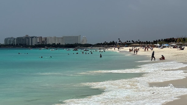 Une plage à l’eau turquoise et limpide, où l’on voit des gens nager et se promener le long du rivage, avec des bâtiments en arrière-plan.