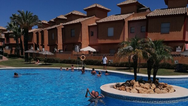 A swimming pool with clear blue water, surrounded by a building with red-tiled roofs and palm trees.