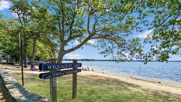 Ein Wegweiser mit der Aufschrift 'Wequaquet Lake' und 'Town of Barnstable' in der Nähe eines Strandes mit Blick auf den See.