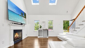 Ein großzügiges Wohnzimmer mit Kamin, weißen Sofas, einem Flachbildfernseher und Blick ins Freie.