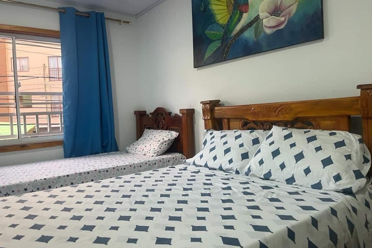 A room with a bed, a window with blue curtains, and a painting of a hummingbird and flower on the wall.