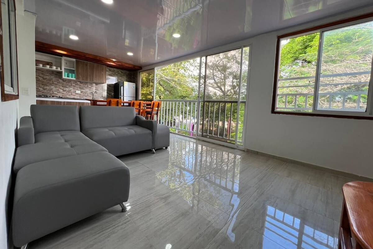 A spacious living area with a grey sectional sofa, a dining table with chairs, a kitchen area, and large windows offering a view of greenery.