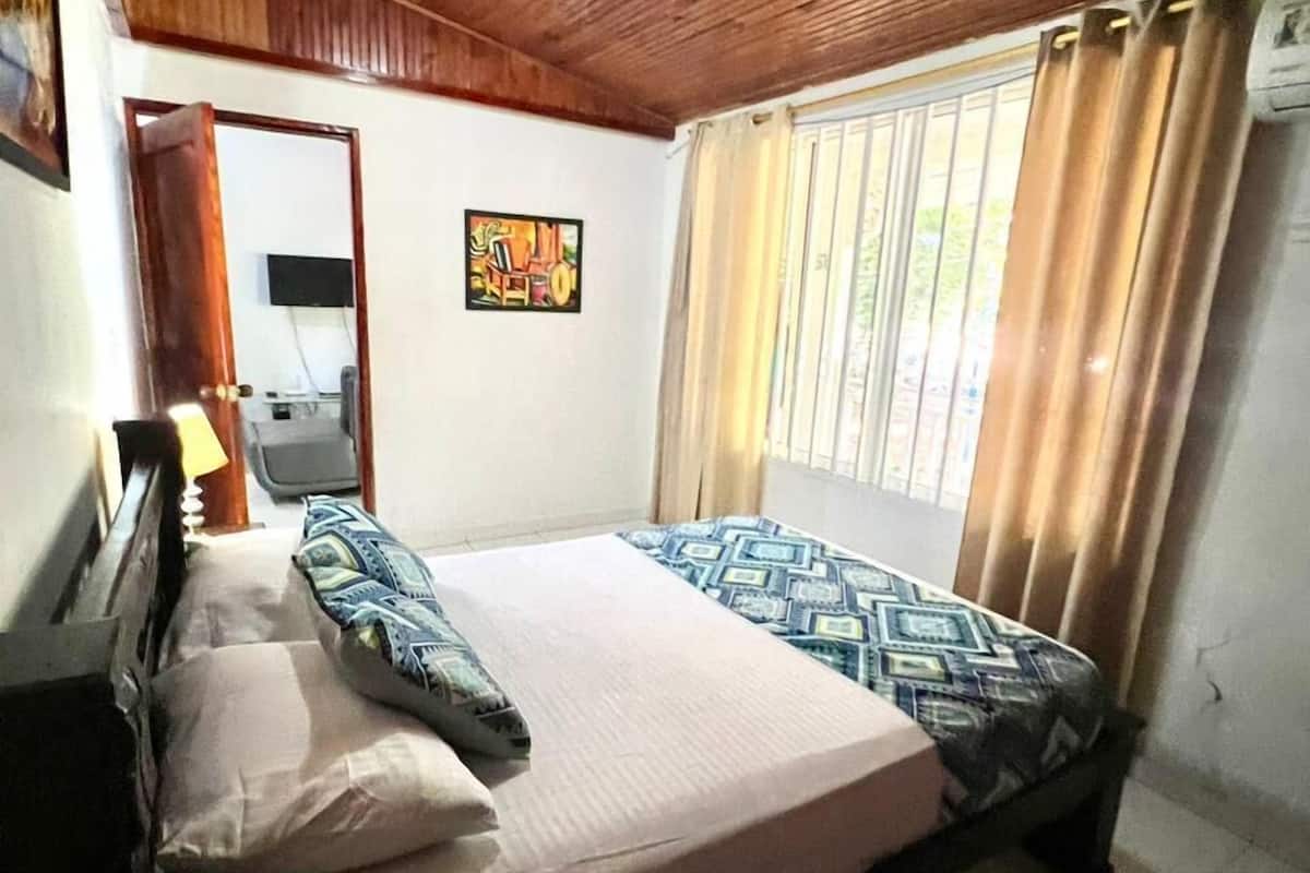 A bedroom with a wooden ceiling, a bed with patterned bedding, a bedside table, a lamp, a window with curtains, and a wall-mounted TV.