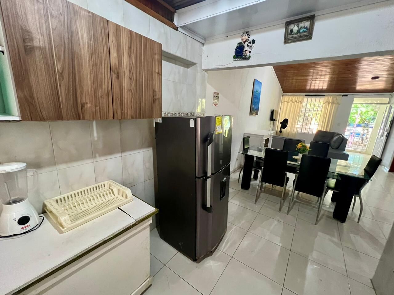 Una cocina con refrigerador, licuadora y lavamanos. Hay una mesa de comedor con sillas y una ventana con cortinas.