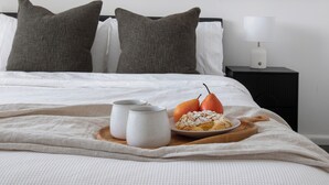 A neatly made bed with a white comforter and gray pillows, a wooden tray with a plate of food, two white mugs, and a single pear. - Enjoy a Luxurious Elwood Escape (Elwood)