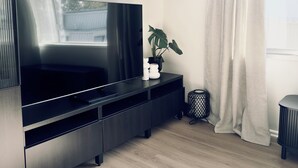 A modern living room with a large flat-screen TV mounted on a dark wooden cabinet, a potted plant on a side table, and a light-colored curtain on a window. - Enjoy a Luxurious Elwood Escape (Elwood)
