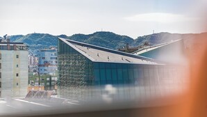 Un edificio moderno con fachada de vidrio y techo inclinado, rodeado de otros edificios y colinas al fondo.