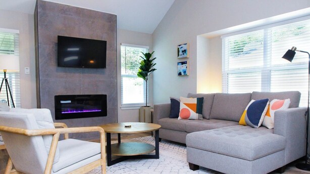 A modern living room with a grey sofa, a wooden chair, a small round table, a flat-screen TV mounted on a wall, and a fireplace.