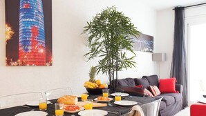 A dining area with a table set for breakfast, including plates, glasses, and a fruit bowl. A large plant and a couch with pillows are in the background.