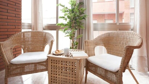 A wicker chair and table set in a bright room with a view of buildings outside.