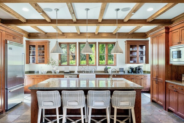 A spacious kitchen with wooden cabinets, a large island with bar stools, pendant lighting, and a view of greenery through large windows.