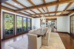 A spacious dining area with a long wooden table, cushioned benches, and large windows offering views of greenery.
