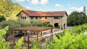 A stone building with a red-tiled roof, a pergola, and a vineyard in the foreground. - Luxury 38-Acre Calistoga Mansion - Pool, Vineyard, Amazing Views, Boccee & More (Calistoga)