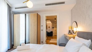 Hotelkamer met een groot bed, nachtkastje met lamp, spiegel en uitzicht op een gang met een deur naar een andere kamer.
