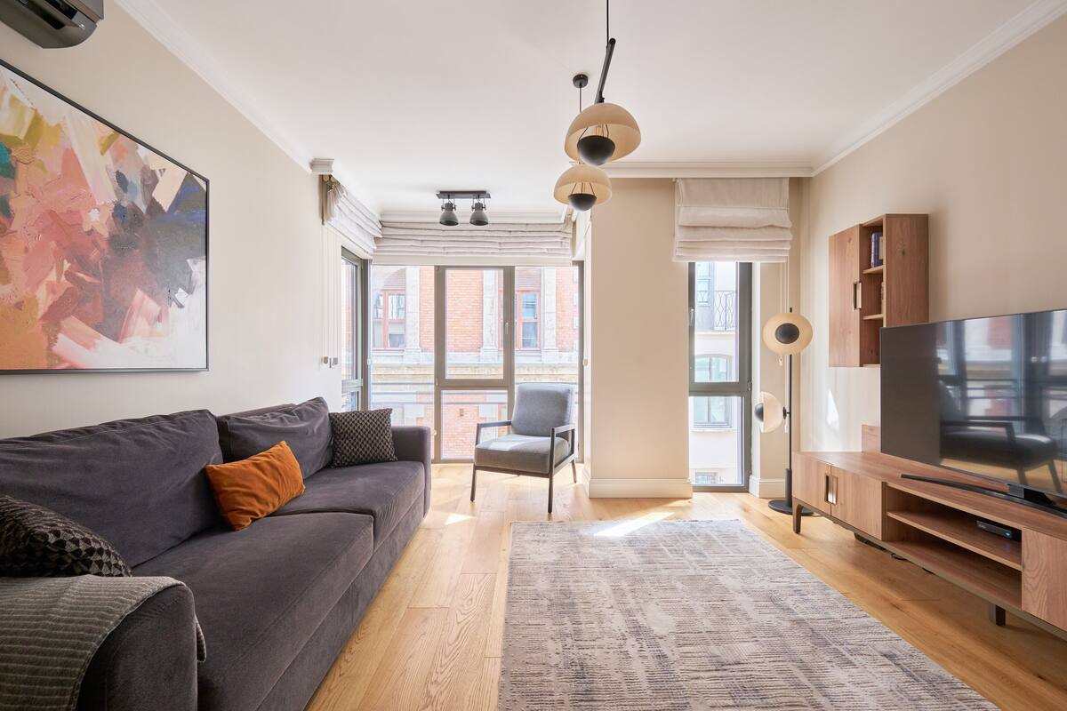 Una sala de estar moderna con un sofá gris, un televisor de pantalla plana, un mueble de madera y un ventanal con vistas a edificios.