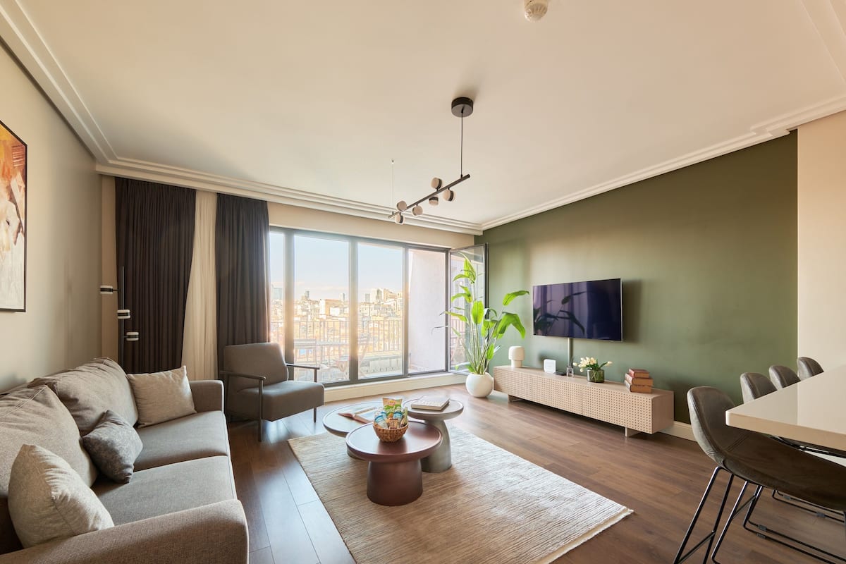 Una sala de estar moderna con sofá, mesa de centro y comedor, ventanas amplias con vista a la ciudad y un televisor de pantalla plana montado en la pared.