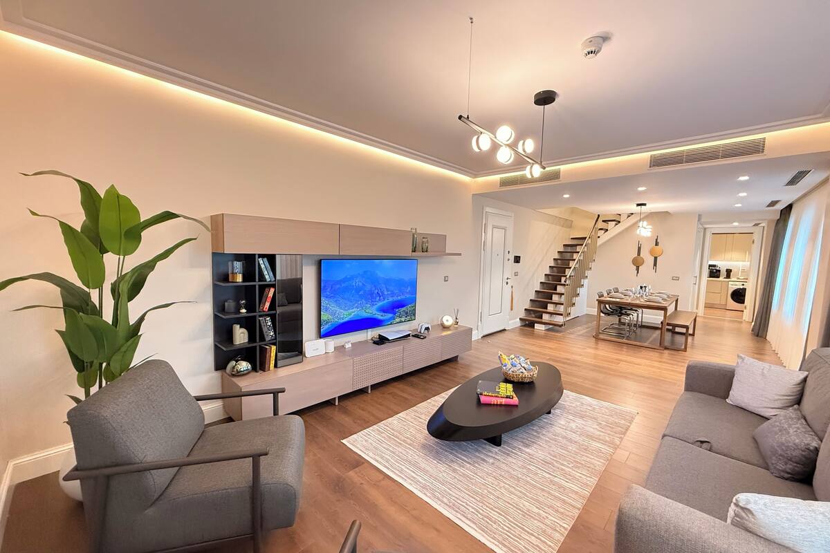 Una sala de estar moderna con un televisor de pantalla plana, una estantería, una mesa de centro y una escalera que lleva a un comedor.