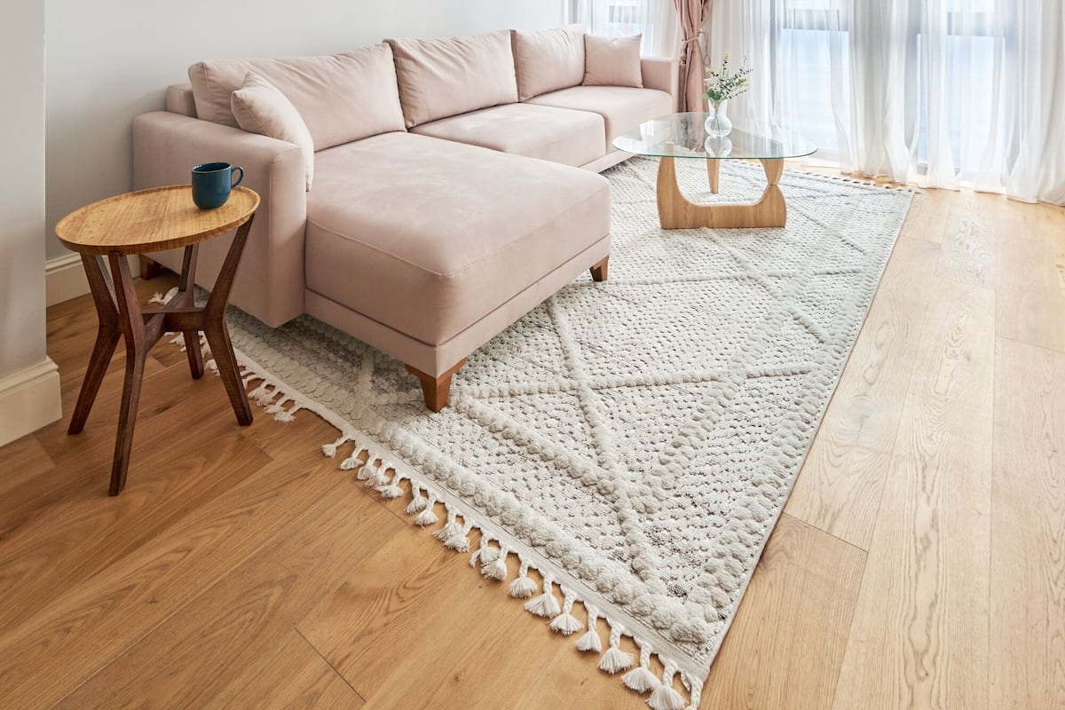 Una sala de estar moderna con un sofá color beige, una mesa de centro de vidrio, una mesita auxiliar de madera y una alfombra blanca de punto.