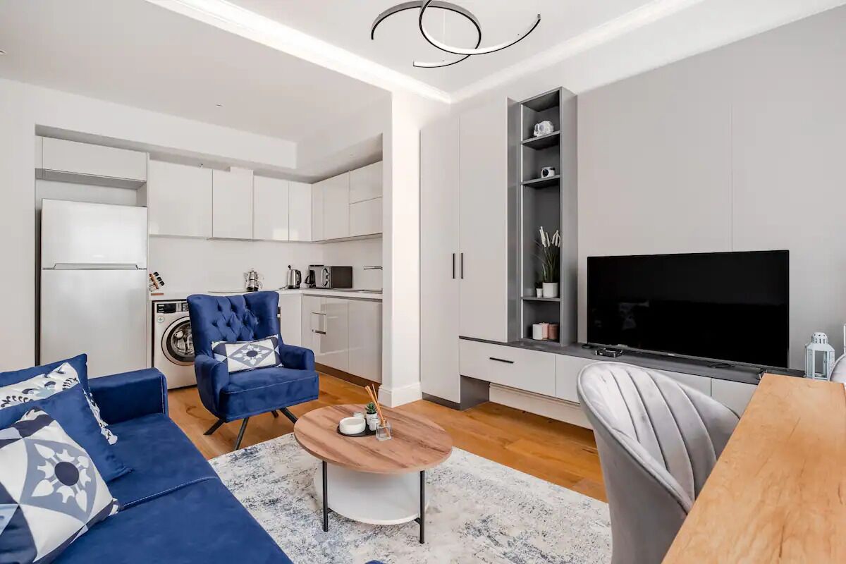 Ein modernes Wohnzimmer mit Sofa, Sessel und Couchtisch. Im Hintergrund ist eine Küche mit weißen Schränken und Geräten zu sehen.
