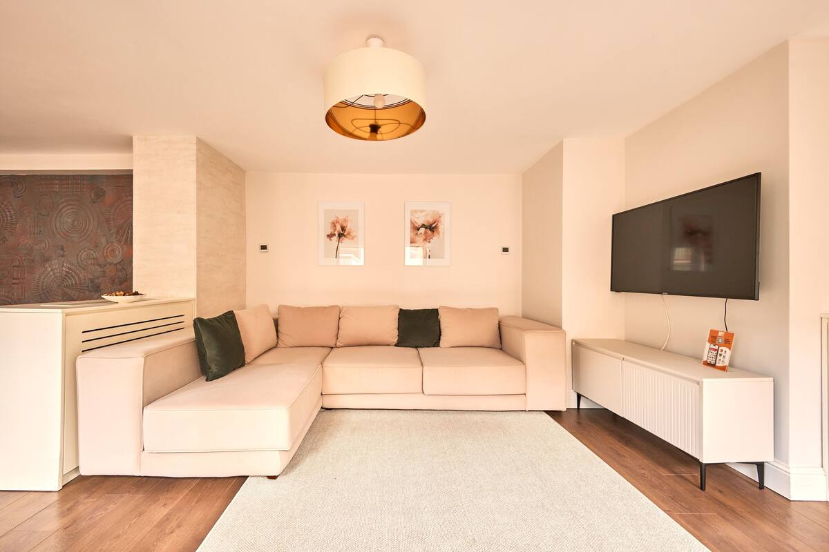 Una sala de estar moderna con un sofá seccional color beige, un radiador blanco, un televisor de pantalla plana y dos obras de arte florales enmarcadas en la pared.