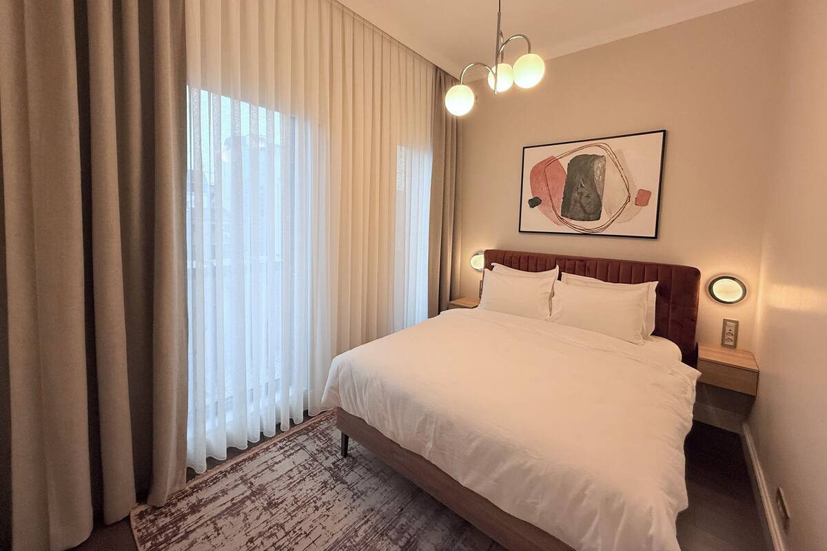 Habitación de hotel con una cama grande, una mesita de noche, una lámpara de pared y un ventanal con cortinas.