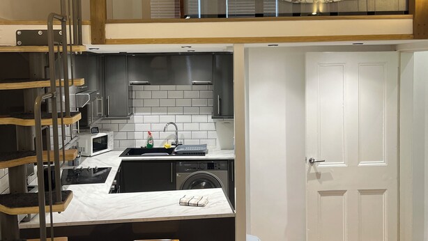 A compact kitchen with a built-in microwave, sink, and a washing machine. There is a small island with stools, a staircase, and a door leading to another room.