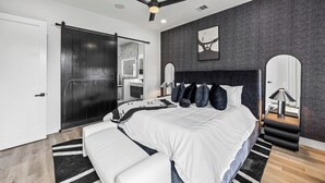 Une chambre moderne avec un grand lit, un ventilateur de plafond, une armoire en bois foncé et une salle de bain visible à travers une porte ouverte.