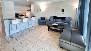 Ein modernes Wohnzimmer mit grauem Sofa, einem Couchtisch und einer Küchenzeile mit weißen Schränken und einer gefliesten Rückwand.