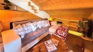 A cozy attic room with a wooden ceiling, a sofa with decorative pillows, a small table with a book, and a staircase leading to the upper floor.