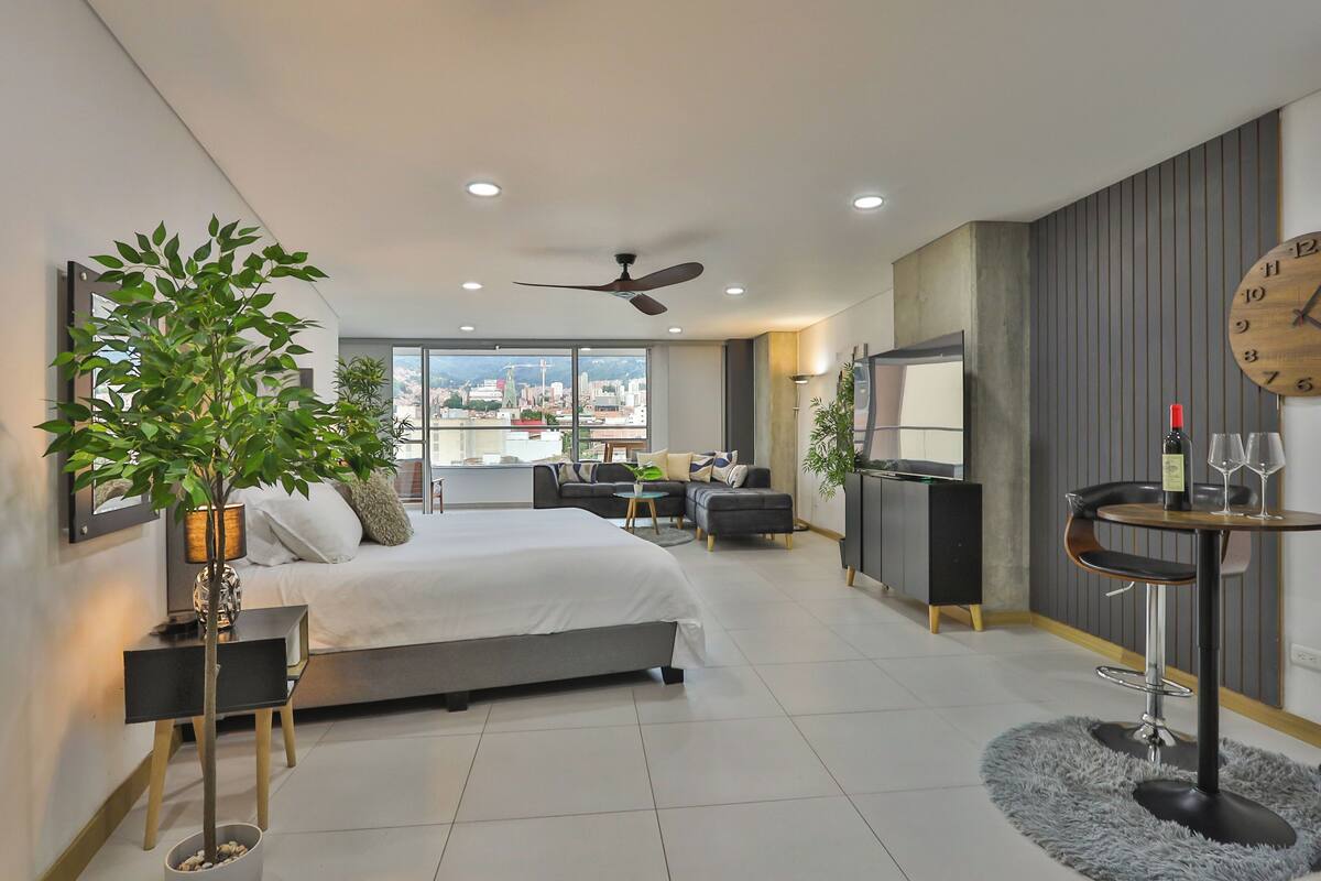 A modern hotel room with a large bed, a dining area with a round table, a TV, and a city view.
