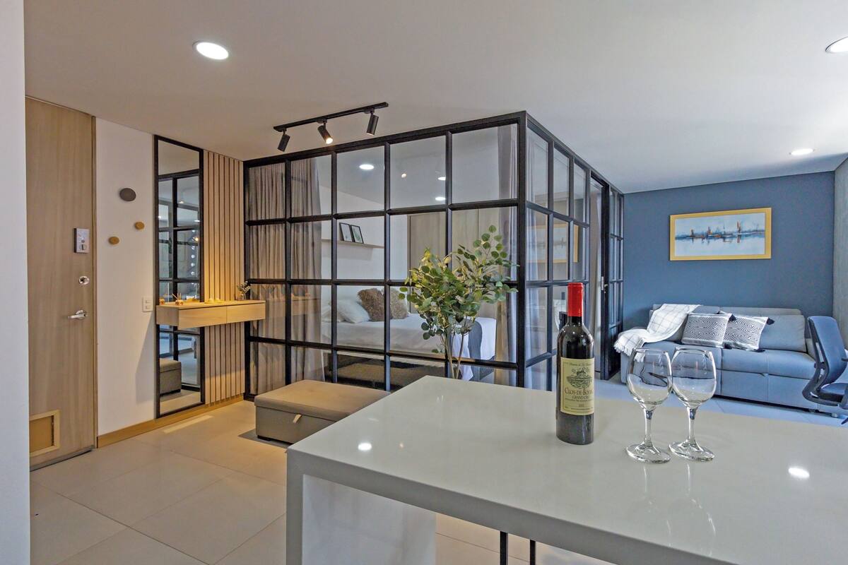 A modern living room with a glass partition, a white table with wine glasses, a sofa, and a painting on the wall.