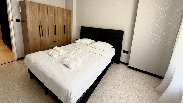 Cama bem arrumada com lençóis brancos e duas toalhas enroladas, guarda-roupa de madeira com duas portas e cabeceira preta.