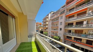 Varanda com grama sintética, guarda-corpo branco e vista para prédios altos com varandas e carros estacionados.