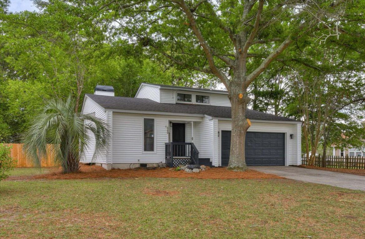 A single-story house with a dark garage door, a front porch with a black railing, and a large tree in front.